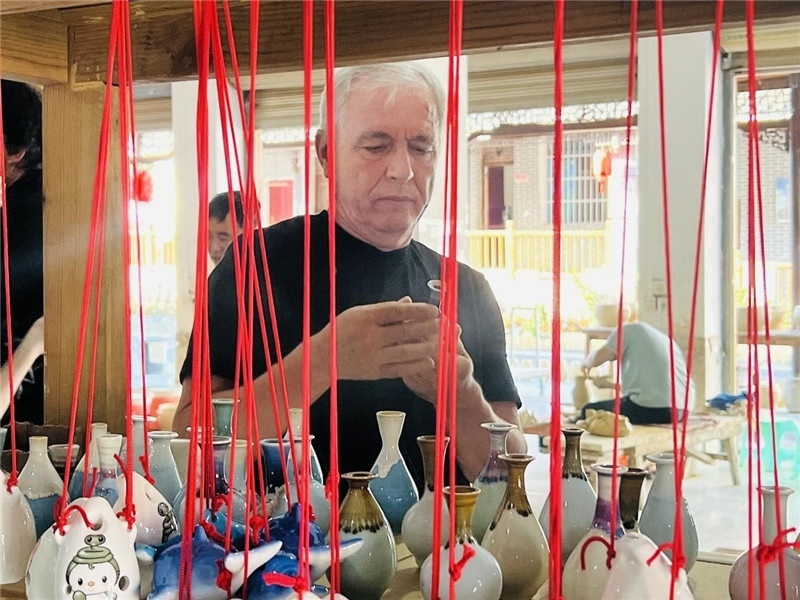 鄉(xiāng)愁如同一根根紅線，將游子與故鄉(xiāng)永遠(yuǎn)地牽連到一起?；逄账囄幕瘎?chuàng)意一條街的母氏陶藝館內(nèi)，紅線吊著陶瓶，思緒游走萬(wàn)里。摩爾多瓦“信息標(biāo)簽”通訊社社長(zhǎng)亞歷山大·塔納斯端詳著館內(nèi)展示的陶藝作品，并挑選出心儀的小擺件帶回摩爾多瓦，也帶走花茂村的“鄉(xiāng)愁”。（張亞欣攝）
